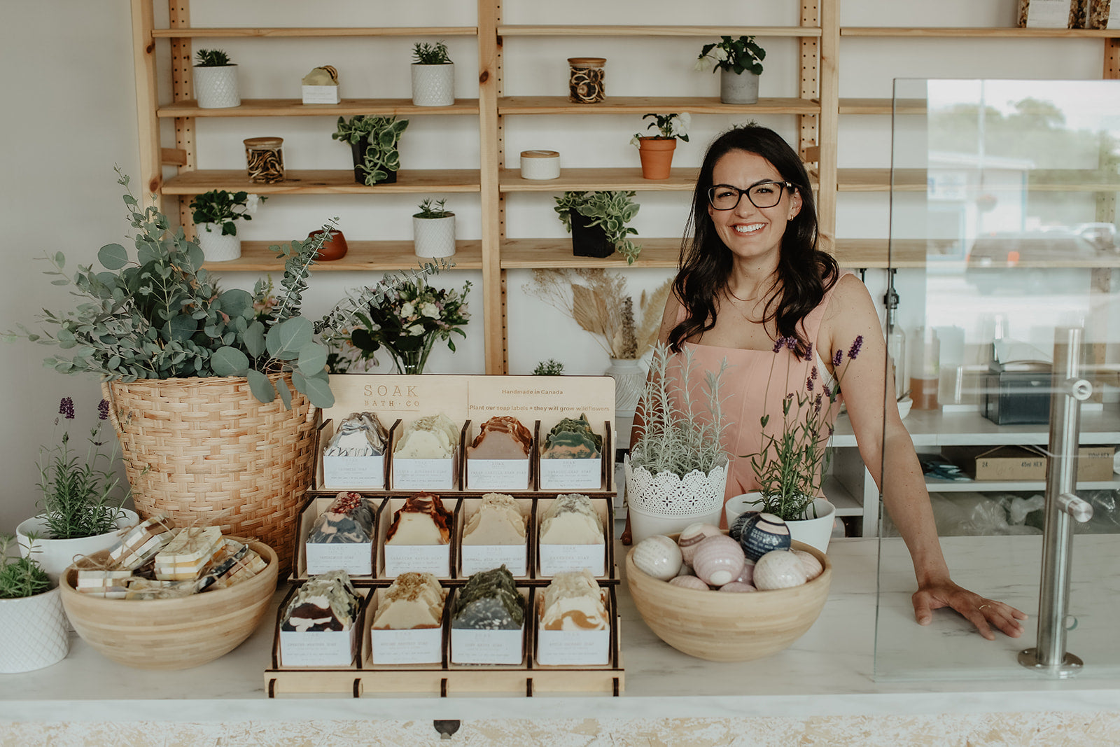 Woman standing behind a counter with various items including plants and soaps, in a store setting.