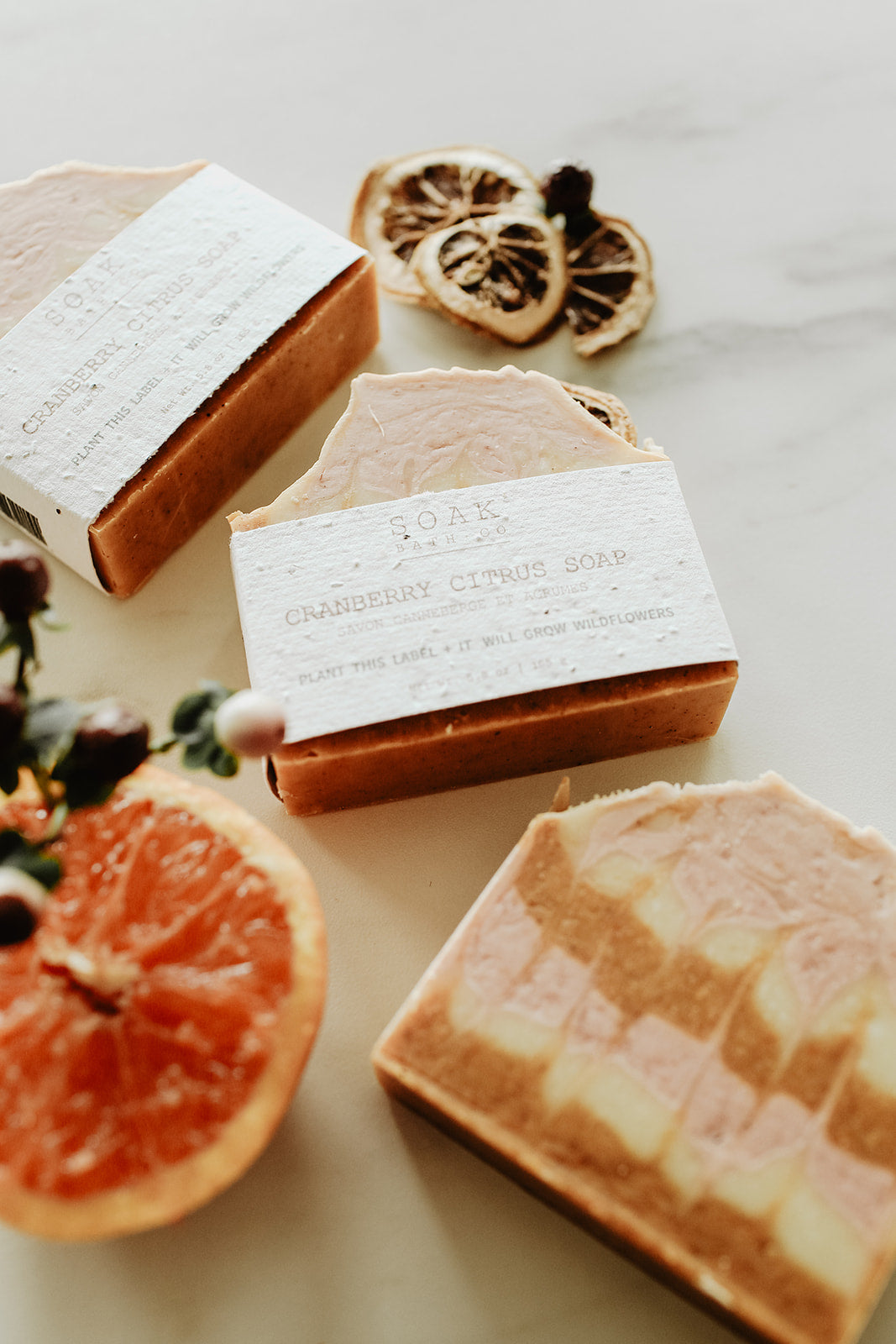 Three soap bars with visible branding on a light surface with dried fruit and citrus.