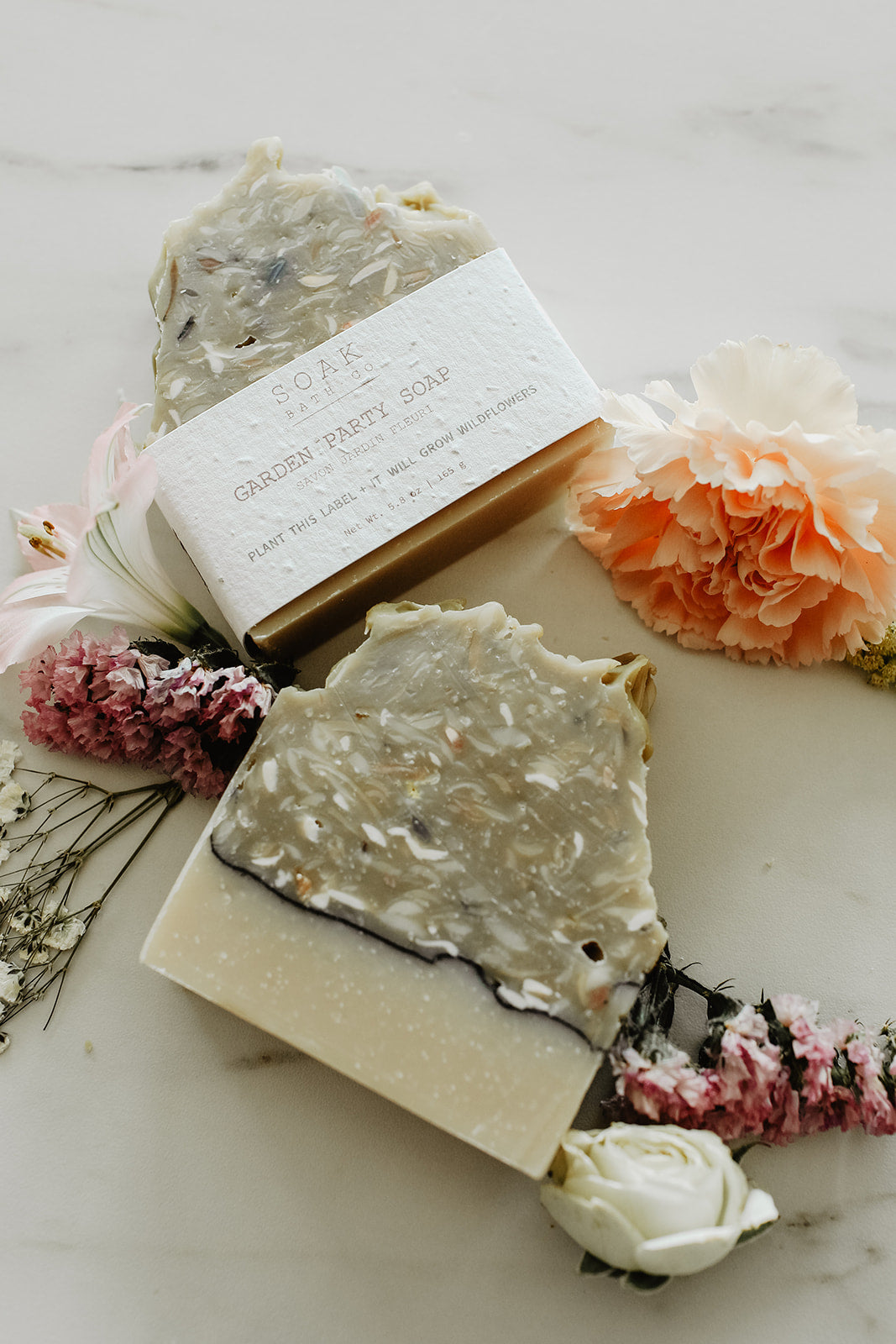 Two bars of soap with a box on a marble surface surrounded by flowers