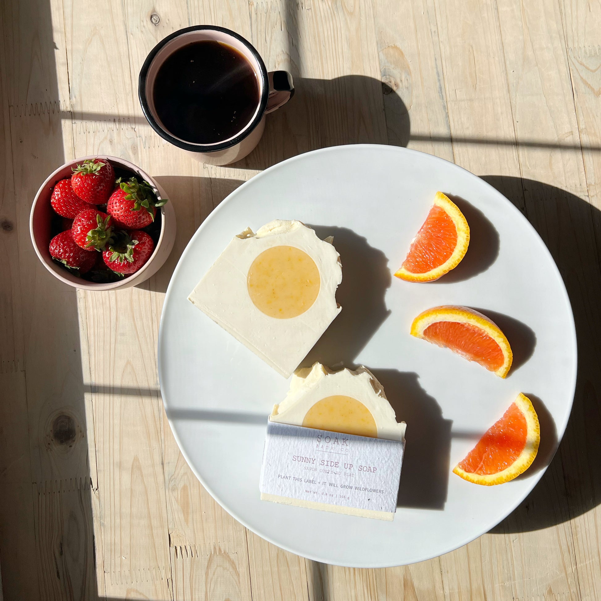 Plate with soap bars, orange wedges, and strawberries next to a cup of coffee on a wooden table.