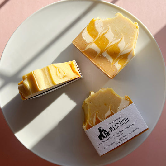 Three artisanal soap bars on a white plate, featuring a yellow and white marbled pattern. One bar is unwrapped, showing its textured top, while another has a white paper label with the brand 'Sib涅pgh Farm Fühls'.