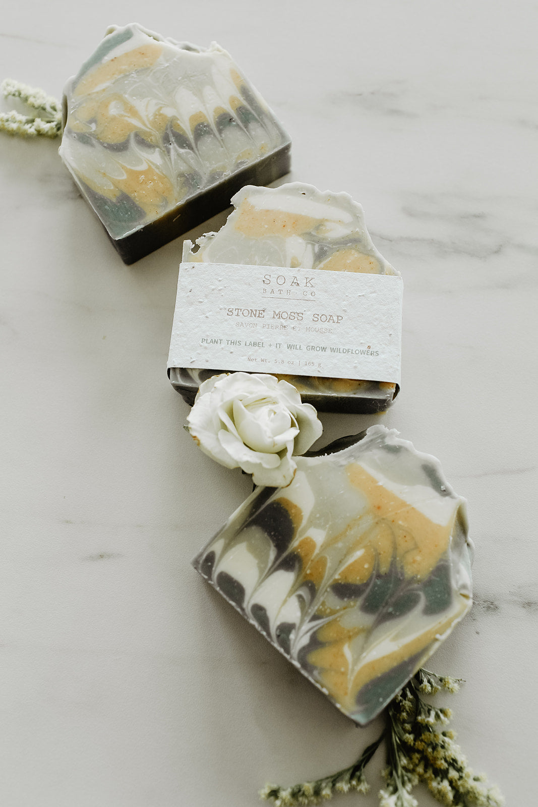 Three hexagonal soap bars with marbled design on a marble surface, accompanied by a white rose and greenery.