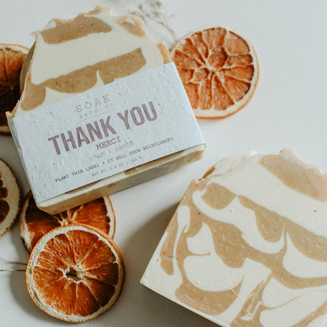 Two soap bars with decorative packaging on a white surface with dried orange slices.