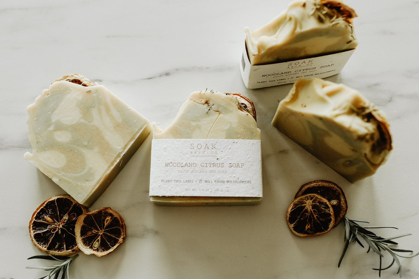 Bar of soap with packaging on a marble surface with dried oranges and rosemary.