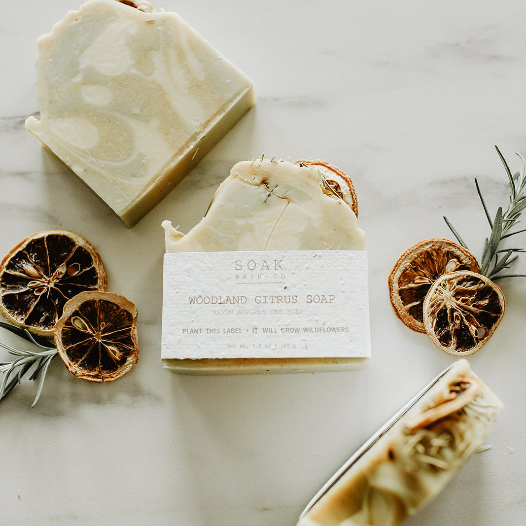 Three bars of soap with a label, surrounded by dried oranges and rosemary on a light surface.
