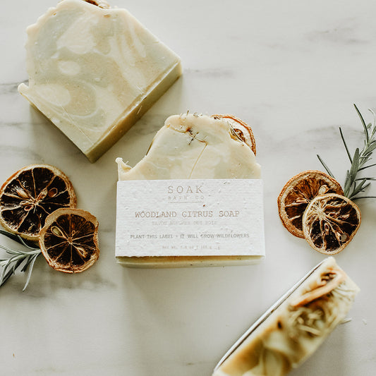 Three bars of soap with a label, surrounded by dried oranges and rosemary on a light surface.